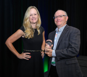 Dr. Beth Keser of Volantis Semiconductor and former IMAPS President stands beside Mike Kelly, who holds an award, both smiling in front of a green-lit backdrop.