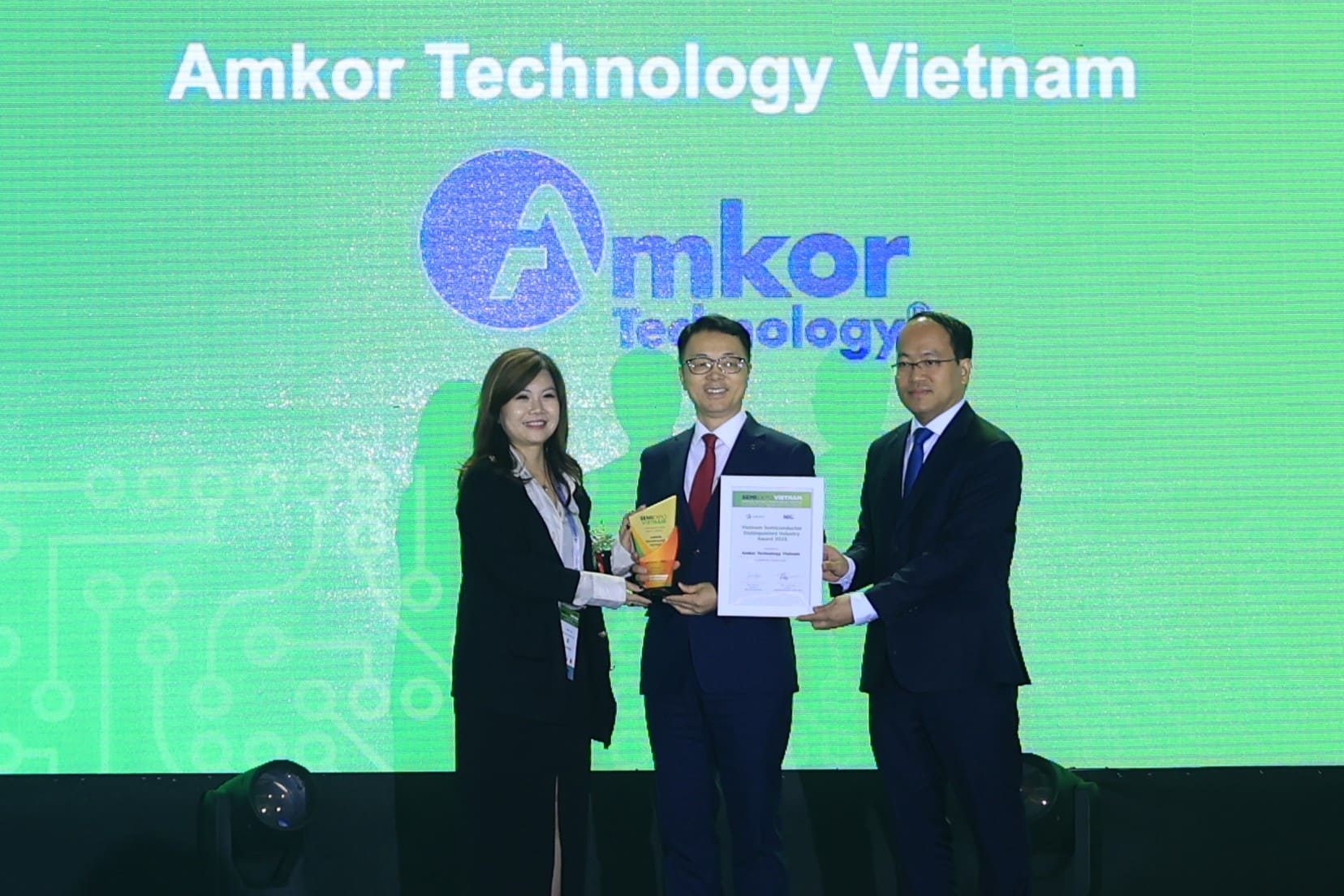 SungHun Kim, Senior Vice President and Country Manager of Amkor Technology Vietnam, stands center stage in formal attire receiving the Vietnam Semiconductor Distinguished Industry Award 2025. He is presented with a plaque and certificate by two presenters, in front of a green display with the Amkor Technology Vietnam logo.