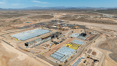 Aerial view of Amkor’s ATA facility under construction in a desert landscape, showing concrete foundations, cranes, construction equipment, work zones, access roads, and surrounding mountains in the distance.
