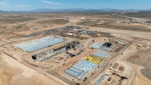 Aerial view of Amkor’s ATA facility under construction in a desert landscape, showing concrete foundations, cranes, construction equipment, work zones, access roads, and surrounding mountains in the distance.