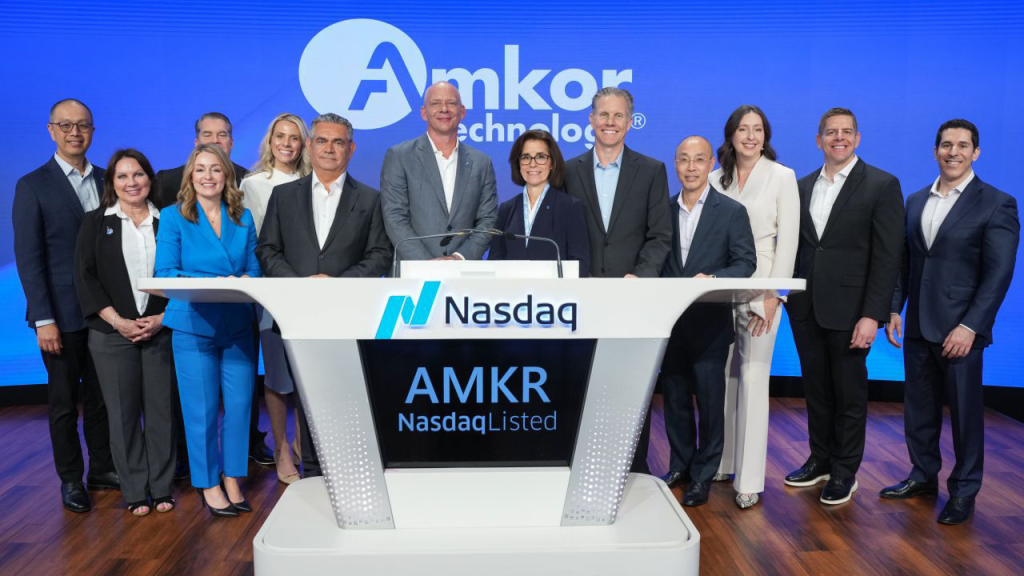 Group of Amkor Technology team members standing behind a Nasdaq podium displaying “AMKR Nasdaq Listed” during a corporate event on a blue branded stage.