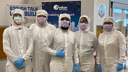 Five individuals dressed in full cleanroom suits, face masks, protective eyewear, and gloves stand together in front of an Amkor Technology display during a semiconductor cleanroom training event.
