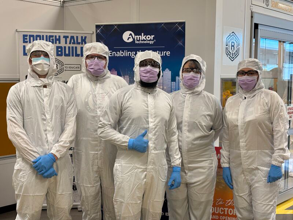 Five individuals dressed in full cleanroom suits, face masks, protective eyewear, and gloves stand together in front of an Amkor Technology display during a semiconductor cleanroom training event.