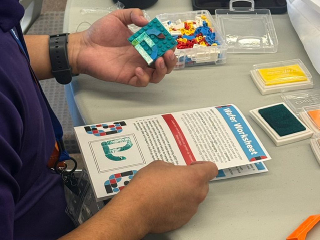 Participant holds a small block-based model while reviewing a “Wafer Worksheet” during a hands-on semiconductor education activity. Colorful building pieces, instructional materials, and workshop supplies are spread across the table.